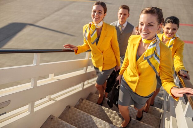 Alegres asistentes de vuelo de hombres y mujeres con uniforme de aviación mientras suben las escaleras de embarque del avión