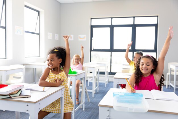 Foto alegres colegialas de primaria levantando la mano mientras están sentadas en el escritorio durante la clase en la escuela