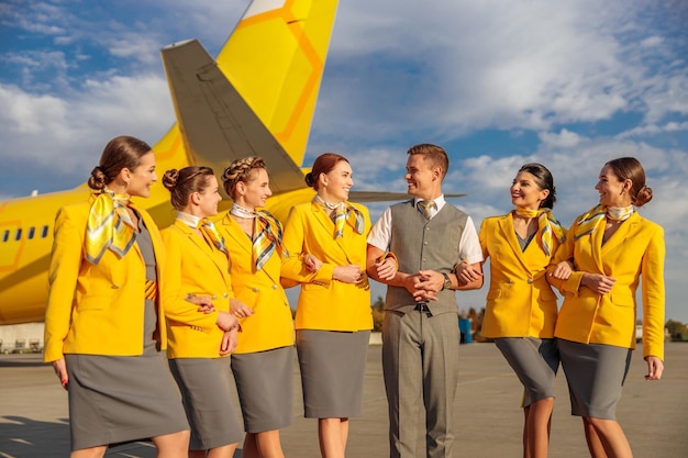 Alegres comissárias de bordo olhando para o colega masculino e sorrindo em pé sob o céu nublado no aeroporto