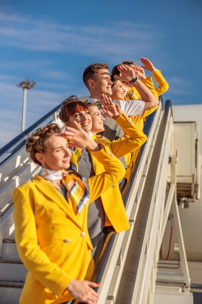 Alegres comissários de bordo de homens e mulheres cobrindo o rosto do sol com a mão e sorrindo em pé nas escadas de embarque do avião