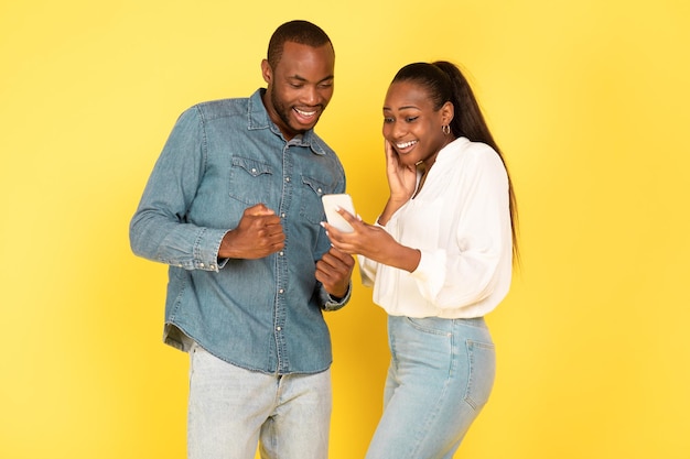 Alegres cônjuges negros usando smartphone agitando os punhos sobre fundo amarelo