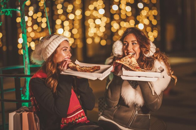 Alegres jóvenes amigas están sentando rebanadas de pizza y divirtiéndose en la noche de invierno en la calle de la ciudad.