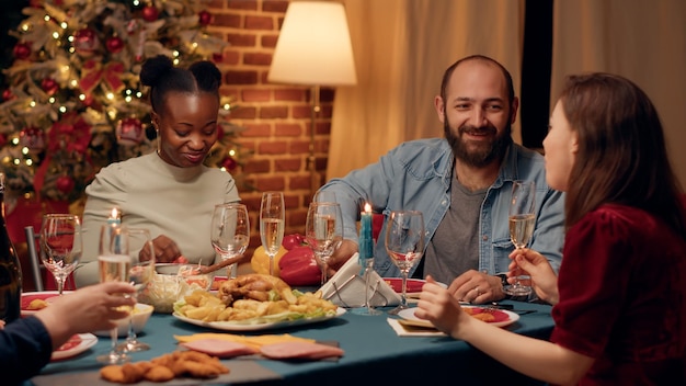 Alegres miembros multiétnicos de la familia brindando champán mientras disfrutan de la cena de Navidad en casa. Gente feliz festiva tintineando copas de vino espumoso mientras celebra las vacaciones de invierno.