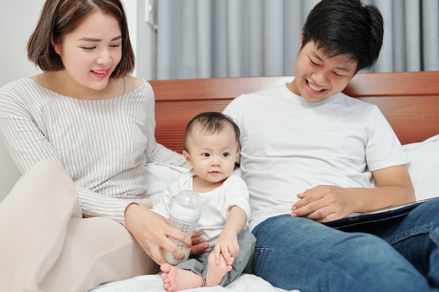 Alegres padres asiáticos jóvenes sentados en la cama, jugando con el pequeño bebé y alimentándola con fórmula