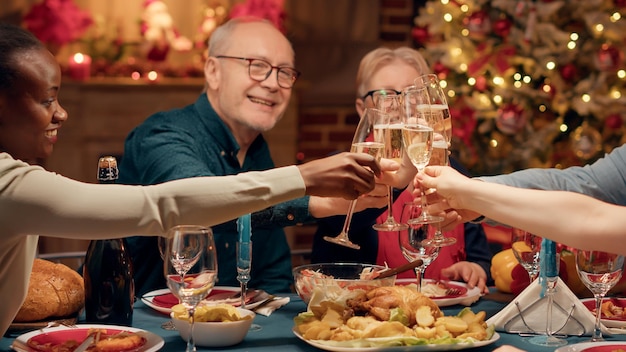 Alegres parentes próximos desfrutando de férias de inverno juntos enquanto bebem vinho espumante. pessoas festivas positivas comemorando o natal no jantar enquanto tilintam copos com champanhe. tiro de tripé