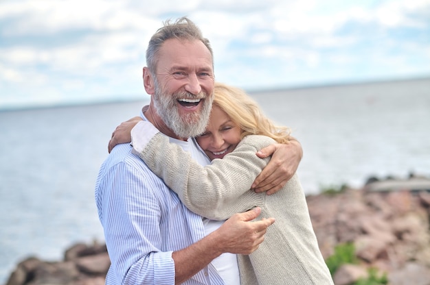 Alegria da vida. homem barbudo adulto encantado abraçando uma fofa mulher sorridente em pé na natureza perto do mar