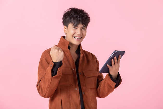 Una alegría exitosa y Expresa Asia un hombre joven y guapo con una tableta a los ojos laterales mirando a la cámara en el amor aislado en el fondo rosado del estudio del espacio de la copia en blanco.