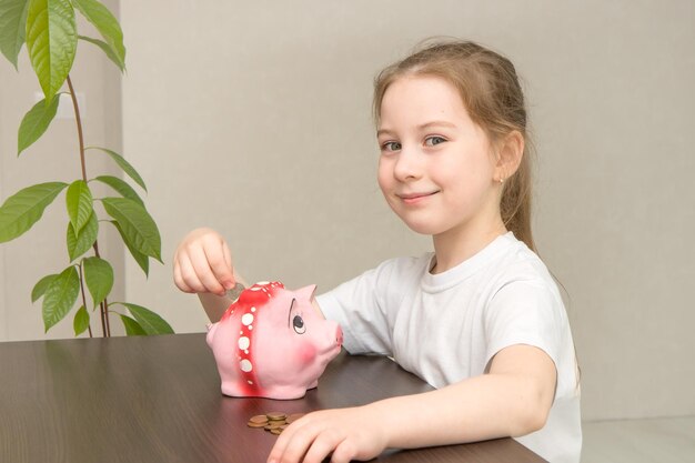 Alfabetização financeira, menina bonitinha sentada à mesa joga moedas no cofrinho e sorri