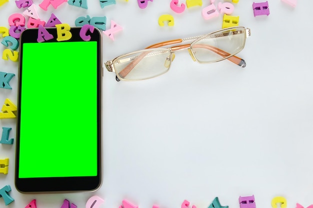 Alfabeto estrangeiro com óculos e um telefone celular com uma tela verde em branco estão sobre um fundo claro
