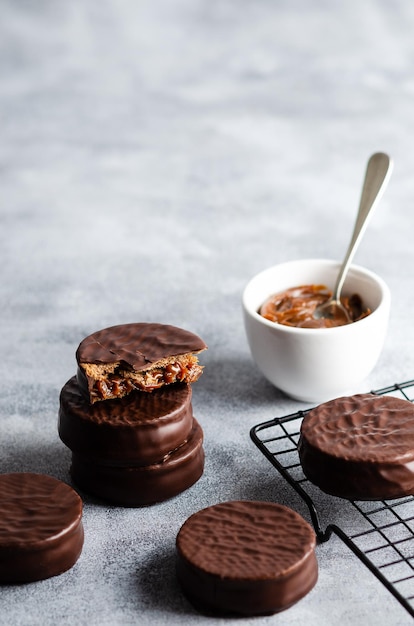 Alfajores de chocolate escuro em um rack escuro e uma tigela com doce de leite em fundo cinza.