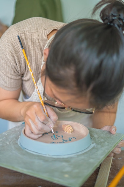 Alfarero de mujer trabajando en la rueda de alfarero haciendo vasija de cerámica de arcilla en el concepto de arte de taller de cerámica Enfoque mano mujer joven adjuntando parte del producto de arcilla al futuro producto de cerámica Taller de cerámica