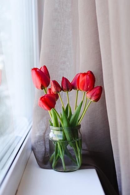 En el alféizar de la ventana cerca de la ventana con una cortina en un frasco de agua hay un ramo de tulipanes rojos