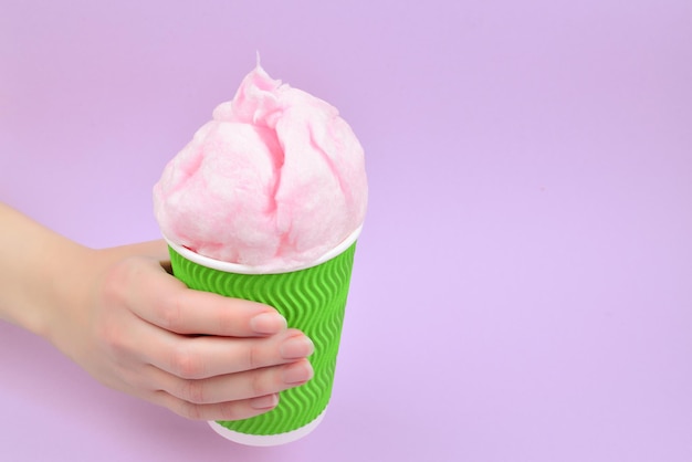 Algodón de azúcar rosa brillante en taza plastick en mano de mujer sobre fondo lila.