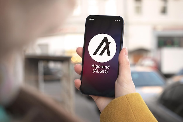 Algorand ALGO Kryptowährungssymbol, Logo. Geschäfts- und Finanzkonzept. Hand mit Smartphone, Bildschirm mit Kryptosymbol-Nahaufnahme