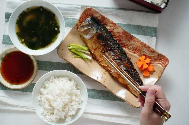 Alguien usando palillos tratando de recoger un saba a la parrilla o pescado caballa servido con sopa de miso y arroz cocido en mantel blanco y verde a rayas en la mesa blanca