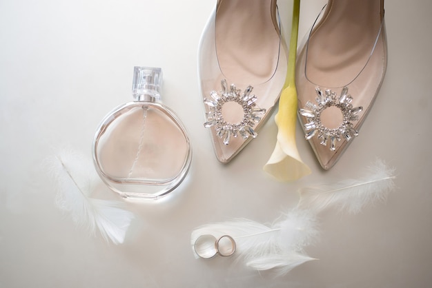 Foto alianças de ouro com penas ao lado dos sapatos bege da noiva decorados com pedras nas quais está uma flor amarela e ao lado de um frasco de perfume chanel