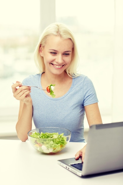 alimentação saudável, dieta, comida, tecnologia e conceito de pessoas - jovem sorridente com computador portátil comendo salada de legumes em casa