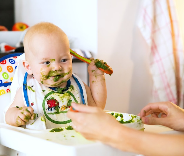 Alimentación Adorable bebé niño comiendo con una cuchara en la silla alta El primer alimento sólido del bebé