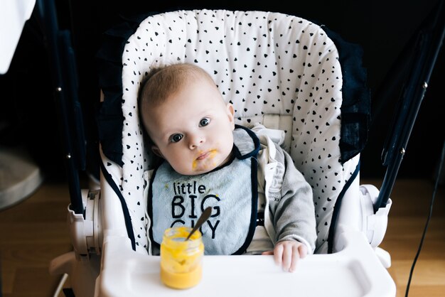 Alimentación. Adorable bebé niño comiendo con una cuchara en silla alta.