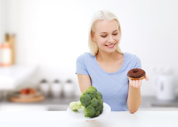 Foto alimentación saludable, comida chatarra, dieta y concepto de elección de personas - mujer sonriente eligiendo entre brócoli y donut sobre fondo de cocina