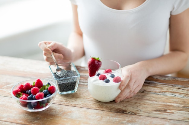 Foto alimentación saludable, comida vegetariana, dieta y concepto de personas - cierre de manos de mujer con yogur, bayas y semillas de amapola o chía en cuchara