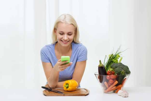 alimentación saludable, comida vegetariana, dieta y concepto de personas - mujer joven sonriente con smartphone cocinando verduras en casa