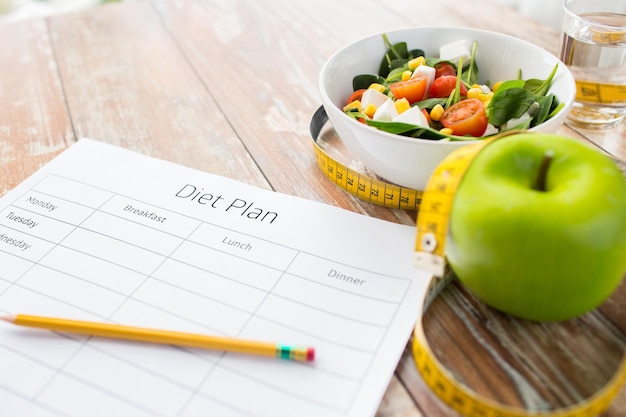 alimentación saludable, dieta, adelgazamiento y concepto de pérdida de peso - primer plano de papel de plan de dieta con manzana verde, cinta métrica y ensalada