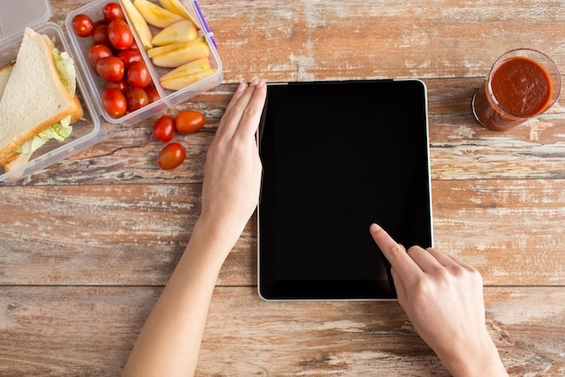alimentación saludable, dieta, tecnología y concepto de personas: cierre de las manos de la mujer con una tableta en blanco y comida en un recipiente de plástico en la mesa en casa