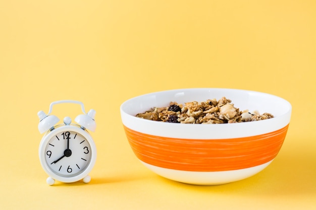 Alimentación saludable. Granola de avena al horno, nueces y pasas en un tazón y reloj despertador sobre fondo amarillo