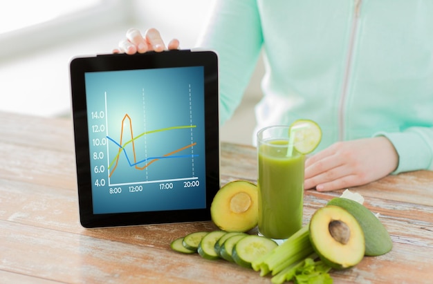 alimentación saludable, tecnología, dieta y concepto de personas: cierre de manos de mujer sosteniendo una computadora de tablet pc con gráfico, jugo verde fresco y verduras sentadas en la mesa