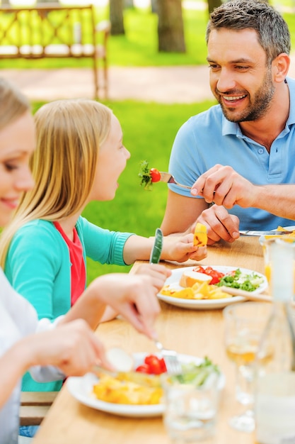 Alimentando con ensalada fresca. Feliz joven alimentando a su hija con ensalada mientras están sentados juntos en la mesa de comedor al aire libre