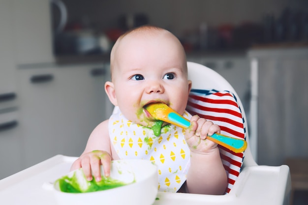 Alimentando um bebê sorridente bagunçado comendo com uma colher na cadeira alta, o primeiro alimento sólido do bebê