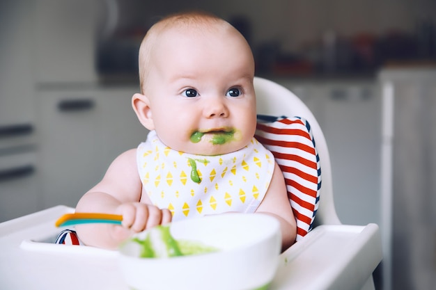 Alimentar al bebé sonriente desordenado comiendo con una cuchara en la silla alta El primer alimento sólido del bebé