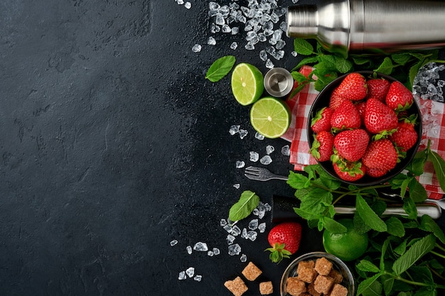 Alimentos ingredientes frescos para hacer limonada, agua de desintoxicación infundida o cóctel. Fresas, lima, menta, albahaca, azúcar de caña, cubitos de hielo y agitador sobre fondo de piedra u hormigón negro. Vista superior.