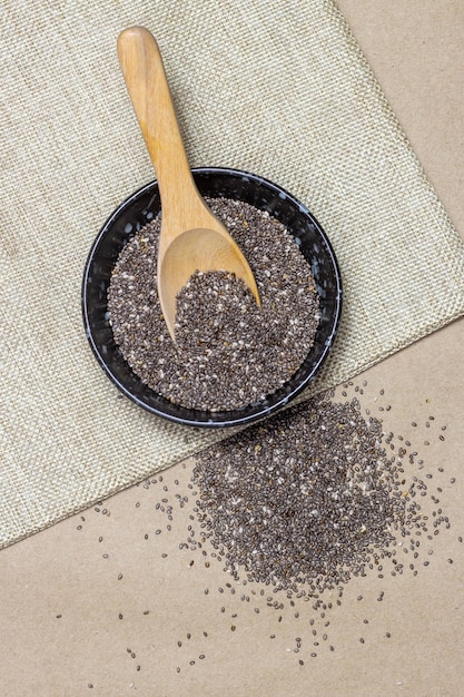 Foto alimentos saudáveis sementes de chia com colher em tigela preta em pano de linho e pilha sobre fundo de papelão