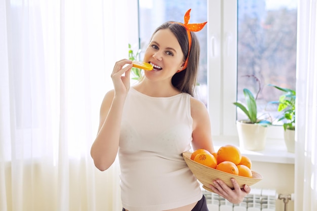 Alimentos saudáveis vitamina, bela jovem grávida feliz com laranjas em casa perto da janela. Gravidez, vitaminas, conceito de maternidade