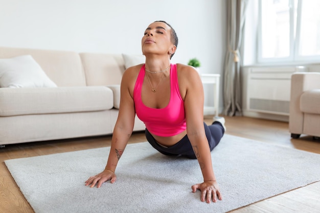 Alivio del dolor de espalda baja mujer negra sonriente haciendo postura de cobra esfinge o asana de perro mirando hacia arriba estirando los músculos de la espalda practicando pilates haciendo ejercicio en el gimnasio o en la sala de estar en la estera de yoga