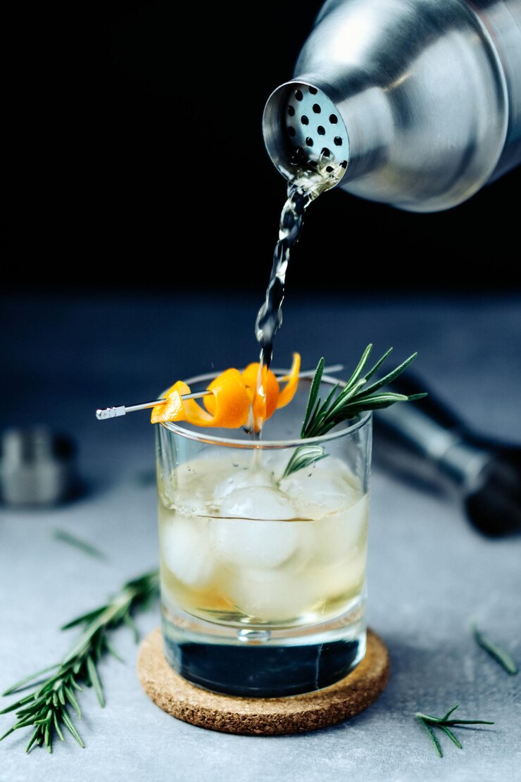 Alkohol-Cocktail aus Shaker in Glas mit Orangenschale, Rosmarin und Eis auf Holzschreibtisch gießen