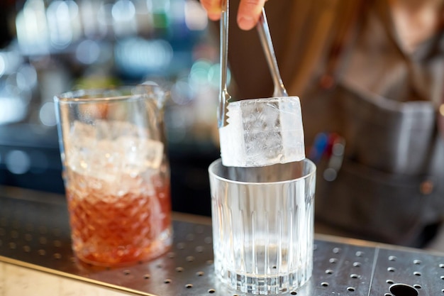 Alkoholgetränke, Menschen und Luxuskonzept - Barkeeper mit Zange, der Eiswürfel ins Glas gibt und Cocktails an der Bartheke zubereitet