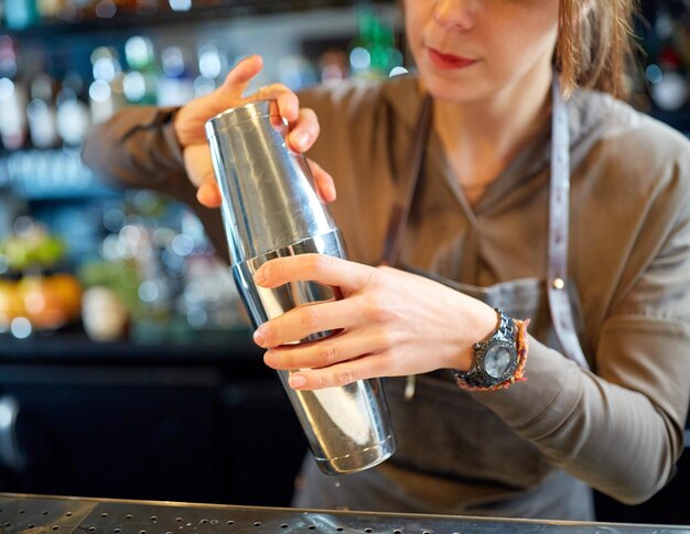 Foto alkoholgetränke, menschen und luxuskonzept - nahaufnahme einer lächelnden barkeeperin mit stahlshaker, die an der bar einen cocktail zubereitet