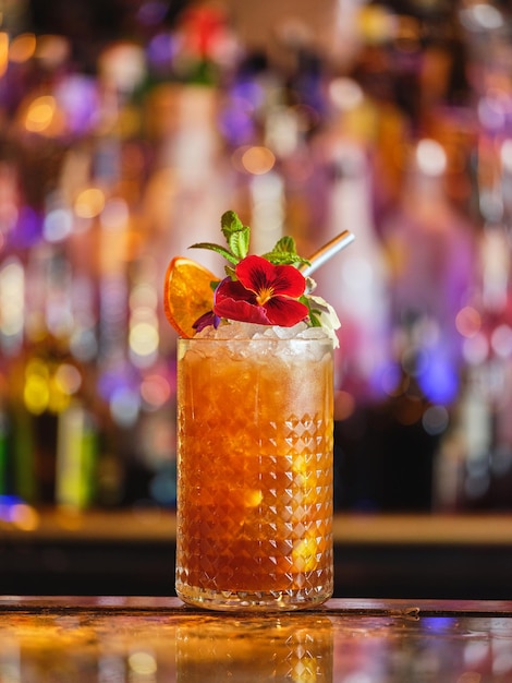 Alkoholischer Cocktail, geschmückt mit essbarer Blume und getrockneter Orange, serviert in einem Glas mit Eiswürfeln in einer Bar auf verschwommenem Hintergrund