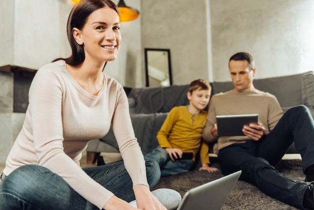 Alle sind beschäftigt. Der Fokus liegt auf einer fröhlichen jungen Frau, die an einem Laptop arbeitet und in die Kamera lächelt, während ihr Mann und ihr Sohn im Hintergrund Videos auf Tablets ansehen