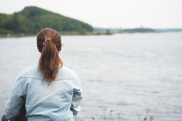 Alleinhippie-Frau, die auf dem Seeufer schaut äußeren Wasser und Gebirgshintergrund sitzt.