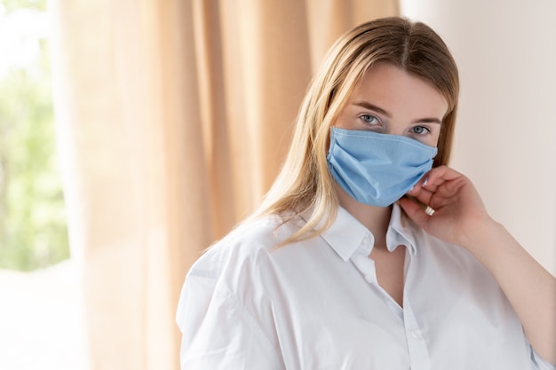 Allergie bei einer jungen schönen Frau. Ein blondes Mädchen in einer medizinischen Schutzmaske steht zu Hause am Fenster und schaut.