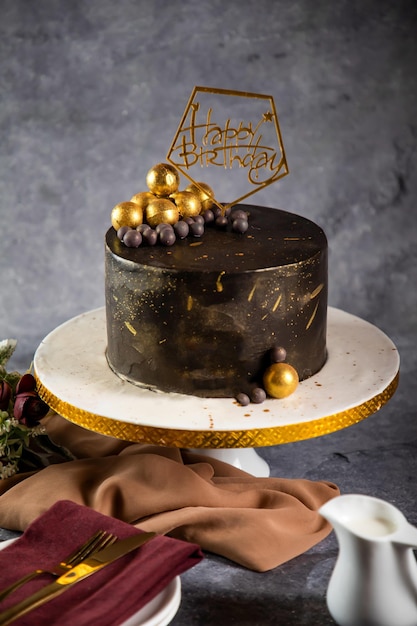 Alles Gute zum Geburtstag-Schokoladenkuchen mit Schokokugeln, Gabel und Messer, serviert in einem Ständer, isoliert auf einer Serviette, Seitenansicht des im Café gebackenen Essens