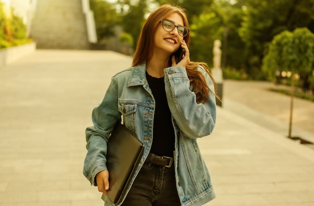 Alles im Geschäft. Junge stilvolle Frau in Jeansjacke und Brille, die Laptop hält und telefoniert, während sie durch die Stadt geht.