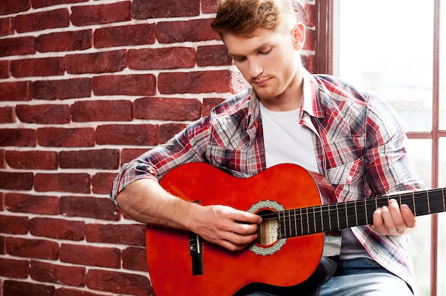 Alma criativa. Jovem bonito tocando violão enquanto está sentado no parapeito da janela