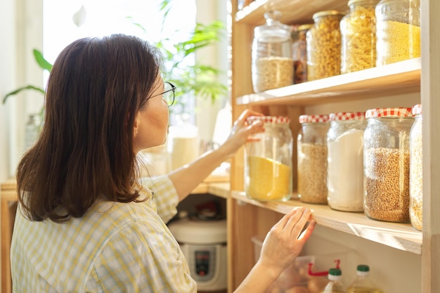 Almacenamiento de alimentos, estante de madera en la despensa con productos de grano en frascos de almacenamiento. Mujer tomando comida para cocinar