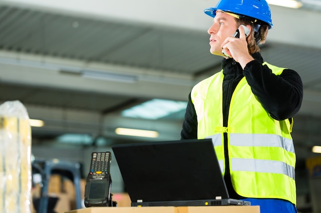 Almacenista con chaleco protector, escáner y computadora portátil en el almacén de la empresa de transporte de carga mediante un teléfono móvil