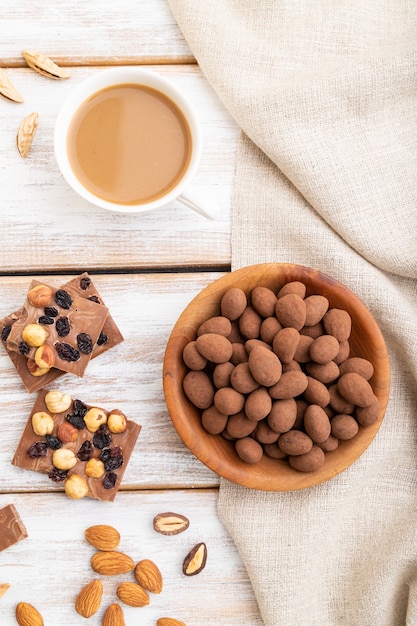 Almendras en grageas de chocolate en placa de madera y una taza de café en la superficie de madera blanca y textiles de lino